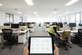 Open-plan office with mixed workstations and a tablet showing a furniture audit interface with red, yellow, and green condition status dots