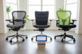 Three iconic task chairs in a GLSC showroom — Herman Miller Aeron, Embody, and Knoll Generation — with a tablet displaying a guided chair selector interface recommending the Embody